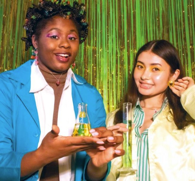 two women hold beakers in front of a neon green background. they are wearing jewelry made out of biodegradable plastic.